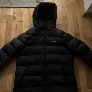 Svart dunjacka Peak Performance DOWN HOOD W - Säljer en svart dunjacka från Peak Performance i storlek XL. i nyskick, används ej pga för stor, nypris 4199kr. En värmande vinterjacka för dam från Peak Performance. Denna jacka är tillverkad i ett tåligt material med en PFC-fri, vattenavvisande ytbehandling och vadderad med förstklassigt ankdun som gör den lätt att packa med sig och passar perfekt till alla utomhusaktiviteter.  Dess höga krage och huva ger extra skydd mot kyliga vindar. Jackan har även två sidofick