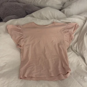 Ljusrosa volangtopp från Kappahl - Supersöt ljusrosa topp från Kappahl med volangdetaljer på ärmarna. T-shirten har rund halsringning och är tillverkad i mjuk bomull. Perfekt för dig som gillar en feminin och chill stil.