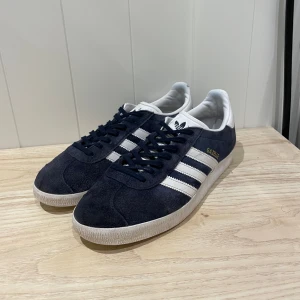 Adidas Gazelle mörkblå mockasneakers - Tjena säljer ett par Adidas Gazelle sneakers i mörkblå mocka med vita detaljer. Skorna har använts väl men har hållits i gott skick. Bara ett par fåtal slitningar (syns i bilden). Skick 7/10. Tveka inte att höra av dig vid funderingar!!
