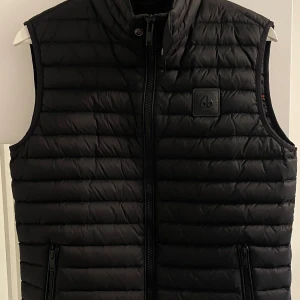 Moose Knuckles Väst - Fin sällsynt svart Moose Knuckles dunväst med quiltad design och hög krage, som ny använd några gånger nypris 3300:- perfekt för alla årstider och alla tillfällen, pris kan diskuteras vid snabb affär.