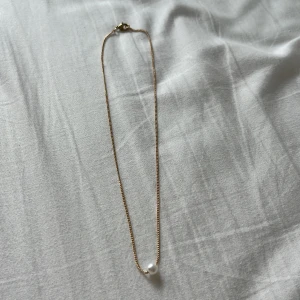 Guldhalsband med pärla - Stilrent halsband med tunn guldfärgad kedja och en ensam vit pärla som hänger i mitten. Perfekt för dig som gillar minimalistiska smycken med en elegant touch.