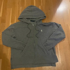 Grå hoodie från Polo Ralph Lauren - Snygg grå hoodie från Polo Ralph Lauren med klassisk vit broderad logga på bröstet. Tröjan har huva med snörning, dragkedja framtill och två fickor. Perfekt för chill dagar och enkel att matcha med jeans eller joggers.Den har en liten skada vid snörrerna fast ingen ser de. Tröjan är rätt liten i storlek, skulle säga passar runt 160-165cm.