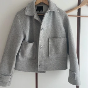 Kappa - Snygg grå jacka i ullblandning från 157. Jackan har en boxig passform, stora framfickor och knäppning med stora knappar framtill. Klassisk krage och breda ärmslut med knappdetalj. Perfekt för lager-på-lager under höst och vår.