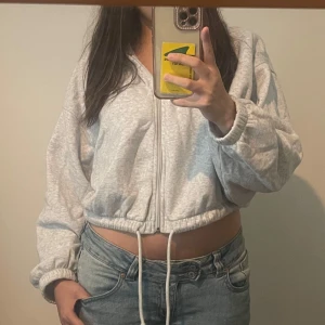 Grå croppad hoodie med snörning, S - Säljer en ljusgrå croppad hoodie med dragkedja och snörning i midjan. Tröjan har långa ärmar med resår vid ärmslut. Mjuk och skön, passar till jeans eller leggings.