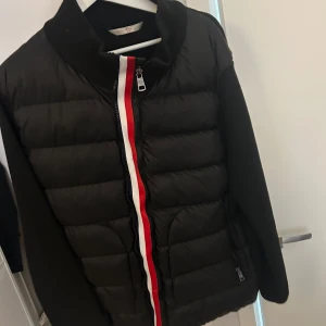 Svart Moncler pufferjacka med röd/vit dragkedja - Snygg svart pufferjacka från Moncler med stickade ärmar och coolt Moncler-märke på armen. Jackan har en dragkedja framtill med röd och vit detalj som ger en sportig vibe. Perfekt för dig som vill ha en stilren och trendig look under kyliga dagar. Xl passar L 185-190 cm