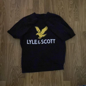 Svart Lyle & Scott sweatshirt med tryck - Svart sweatshirt från Lyle & Scott med stort gult fågeltryck och vit logga på bröstet. Tröjan har rund hals, långa ärmar och ribbade muddar. Perfekt för dig som gillar klassisk streetstyle och vill sticka ut med en ikonisk logga.