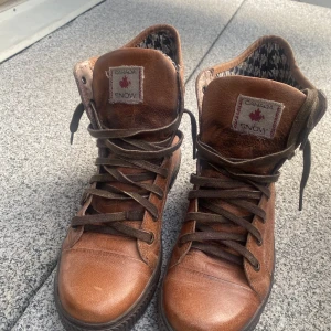 Bruna boots från Canada Snow i skinn - Snygga bruna boots från Canada Snow i äkta skinn med mörkbruna snören och coolt mönstrat innerfoder. Skorna har rund tå, platt sula och högt skaft som ger en urban vibe. Perfekta för dig som vill ha både stil och komfort under kyligare dagar.