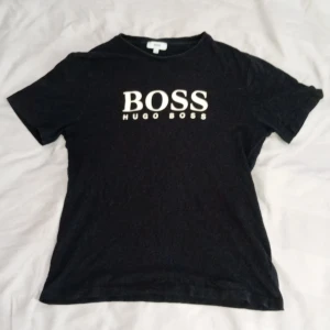 Svart Hugo Boss t-shirt med tryck - Svart t-shirt från Hugo Boss med stort vitt BOSS-tryck på bröstet. Klassisk rund hals och korta ärmar. T-shirten har en normal passform och är gjord i mjuk bomull som känns skön mot huden. Perfekt för dig som gillar stilrena logotoplagg.