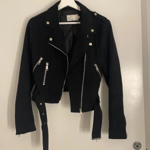 Svart mocka från NLY Trend - Snygg svart mockajacka från NLY Trend med silvriga dragkedjor, knappar och bälte i midjan. Jackan har en croppad passform och långa ärmar. Perfekt för dig som vill ha en edgy look med coola detaljer.