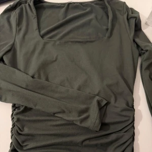 Mörkgrön långärmad topp med rynk - Snygg mörkgrön långärmad topp med fyrkantig ringning och rynkade sidor. Perfekt för dig som gillar stilrena plagg med en twist. Mjuk och stretchig känsla, sitter skönt och tajt på kroppen och passar till många olika looks.