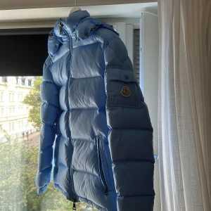 Ljusblå Moncler dunjacka med huva - Säljer en sjukt snygg ljusblå dunjacka från Moncler som är (Limit) med avtagbar luva och dragkedja framtill. Jackan har två fickor med dragkedja, klassisk Moncler-logga på ärmen och coolt serietidningsfoder på insidan. Perfekt för kalla dagar när du vill sticka ut.