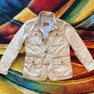 Beige feildjacket från Massimo Dutti  - Stilren beige feildjacket från Massimo Dutti med fyra stora fickor framtill, snygga knappar och dragkedja. Jackan har klassisk krage, markerad midja och detaljerade ärmslut med knappar. Perfekt för dig som gillar en clean och tidlös look. Storlek S ganska så sällsynt skick mycket bra det enda är att den är lite skrynklig men det går o lösa snabbt med ett stryckjärn DM för fler bilder eller frågor 🥯🐕🦤