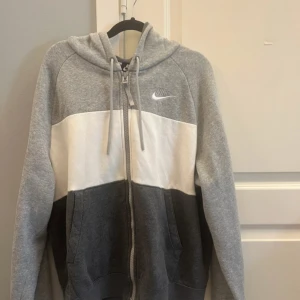Nike Air hoodie i grått och vitt - Nike Air hoodie med dragkedja, huva och snörning. Färgblockad i ljusgrått, vitt och mörkgrått med broderad Nike Air-logga fram och stor tryckt logga bak. Mjuk bomullsmix, två fickor och ribbade muddar. Perfekt för chill eller träning.