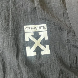 Svart vindjacka från Off-White - Svart vindjacka från Off-White med ikoniska loggor på bröstet och ryggen. Jackan har huva, dragkedja med Off-White-tryck och två fickor framtill. Lätt material som passar perfekt för streetwear-stil.