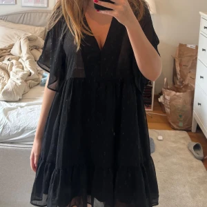 Svart prickig kortklänning från Zara - Säljer en svart kortklänning från Zara med v-ringning och knappar framtill. Klänningen har ett skirt, prickigt tyg och volangdetaljer nedtill. Den är luftig med korta, fladdriga ärmar. Använd 1 gång💕☺️