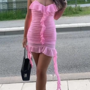 Rosa omlott volangklänning - Supersöt rosa kortklänning med volanger och omlott-detaljer. Klänningen är tight och har offshoulder-design med rynkade detaljer längs hela kroppen. Perfekt för dig som vill sticka ut med en feminin och trendig look.