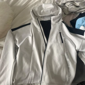 Vit och svart kofta från The North Face - Snygg vit och svart kofta  från The North Face med huva och dragkedja. Koftan  har logga tryckt längs ena sidan och på ryggen, samt svarta detaljer på axlar och rygg. Perfekt för dig som gillar sportig och clean stil.