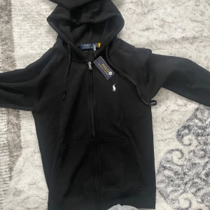 Svart hoodie från Polo Ralph Lauren - Svart hoodie från Polo Ralph Lauren med klassisk vit broderad logga på bröstet. Tröjan har dragkedja, huva med snörning och två fickor framtill. Tillverkad i mjuk bomull, perfekt för en chill och stilren look.