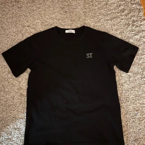 Svart t-shirt från Saint Levitica - Svart t-shirt från Saint Levitica med vit ST-logga på bröstet och stor text på ryggen. Klassisk passform med rund hals och korta ärmar. Skön och stilren, perfekt till jeans eller shorts.