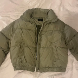 Olivgrön pufferjacka med dragkedja - Säljer en olivgrön pufferjacka med hög krage och dragkedja framtill. Jackan har två fickor på sidorna och elastisk nederkant. Den är quiltad och har en loose passform, perfekt för kalla dagar. Enkel och stilren design som är lätt att matcha.