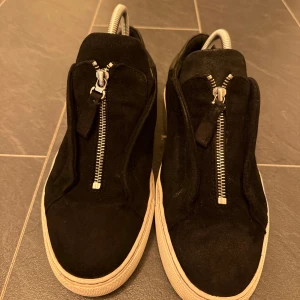 Svarta sneakers från Axel Arigato - Stilrena svarta sneakers från Axel Arigato i mocka med vit platt sula. Skorna har en unik dragkedja framtill istället för klassisk snörning och diskret logga på sidan. Perfekta för dig som gillar minimalistisk och modern design.