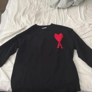 Svart stickad tröja Ami Paris - Svart stickad tröja från Ami Paris med röd hjärt- och A-logga på bröstet. Klassisk rund halsringning och ribbade muddar. Perfekt för dig som gillar stilrena plagg med en twist. Materialet är mjukt och passar till lager-på-lager.