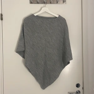 Grå stickad ponchotröja - Säljer en grå stickad ponchotröja med lös passform och asymmetrisk nederkant från Glitter. Perfekt att slänga över en t-shirt eller långärmad tröja för extra värme. Enkel och stilren design som passar till många outfits. (Aldrig använd)