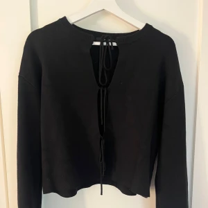 Svart kofta med snörning från Gina Tricot - Svart kofta från Gina Tricot med lång ärm och trendig knyt stängning framtill. Modellen är croppad och har en rak passform. Perfekt att styla med höga jeans eller kjol för en cool look. Mycket bra skick.