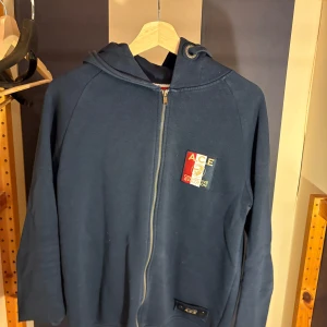 Mörkblå hoodie från A.C.E med dragkedja - Säljer en mörkblå hoodie från A.C.E med hel dragkedja och huva. Tröjan har ett broderat märke på bröstet i rött, vitt och blått samt snörning i huvan med snygga detaljer. Perfekt för dig som gillar streetwear och vill ha en stilren look. Storlek Small men mer som en medium
