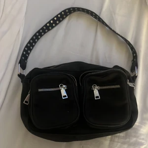 Svart axelväska med nitar och dragkedjor - Cool svart axelväska med bred axelrem dekorerad med silvriga nitar. Väskan har två stora framfickor med dragkedjor och en större dragkedja upptill. Materialet känns som mocka och detaljerna i silver ger en edgy vibe.