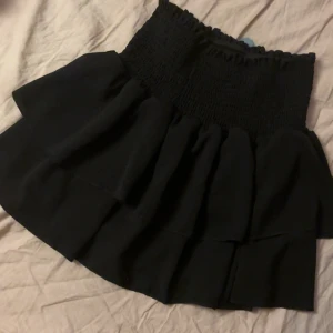 Svart volangkjol med smock - Supersöt svart kortkjol med bred smock i midjan och flera lager volanger. Kjolen har en fluffig och trendig look som passar perfekt till sneakers eller boots. Materialet känns mjukt och följsamt, och midjan är elastisk för extra komfort.