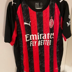 AC Milan fotbollströja Puma röd/svart - AC Milan matchtröja från Puma med klassiska röda och svarta vertikala ränder, klubbmärke på bröstet och vit Emirates-tryck. Kortärmad modell i lätt, ventilerande material. Snygga detaljer på ärmar och axlar, perfekt för dig som vill representera Milan.