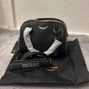 Svart Zadig & Voltaire axelväska i skinn - Cool svart axelväska från Zadig & Voltaire i grymt snyggt skinn med silverfärgade nitar och dragkedjor. Väskan har ett emblem med vingar framtill och bred axelrem med broderad logga. Insidan har ett mönstrat foder i svartvitt.