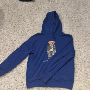 Blå Polo Bear hoodie från Ralph Lauren - Säljer en blå hoodie från Ralph Lauren med ikoniska Polo Bear-trycket på bröstet. Tröjan har huva med snörning och långa ärmar. Perfekt för dig som vill ha en klassisk streetstyle-look med en twist. Materialet är mjuk bomull som känns skönt mot huden.