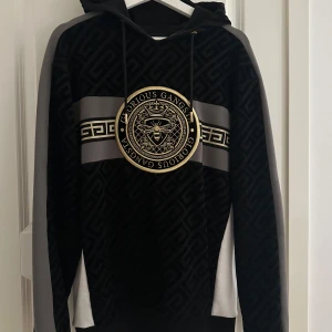 Svart hoodie från Glorious Gangsta - Säljer en svart hoodie från Glorious Gangsta med guldigt emblem och mönstrad struktur. Tröjan har huva med snörning, gråa och vita detaljer på ärmarna samt ett stort tryck framtill. Perfekt för dig som vill sticka ut med en streetstyle-look.