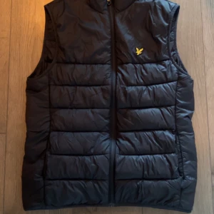 Svart dunväst från Lyle & Scott - Säljer en svart dunväst från Lyle & Scott med klassisk gul logga på bröstet. Västen är quiltad och har dragkedja framtill. Perfekt att slänga över en hoodie eller tröja för extra värme. Enkel och stilren design som funkar till det mesta.