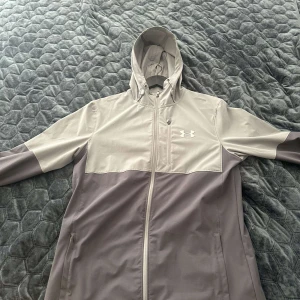 Under Armour vindjacka i grått - Snygg tvåfärgad vindjacka från Under Armour i ljusgrå och mörkgrå nyans. Jackan har huva med snörning, dragkedja framtill och praktiska fickor. Perfekt för dig som gillar sportig stil och vill ha något lätt och funktionellt.