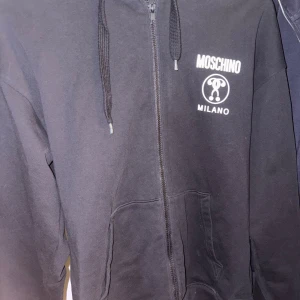 Svart Moschino hoodie med logga - Svart hoodie från Moschino med vit logga och Milano-tryck på bröstet. Dragkedja framtill, huva med snörning och fickor. Snygga vita band med Moschino-symbol längs ärmarna. Perfekt för dig som gillar streetwear och vill sticka ut med designerplagg.