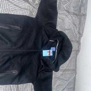 Svart NOCTA x Nike vindjacka med huva - Svart vindjacka från Nike NOCTA med huva och dragkedja framtill. Jackan har snygga sömdetaljer, NOCTA-logga i gult på ärmen och diskreta broderade detaljer. Perfekt för dig som vill ha en clean och sportig look.