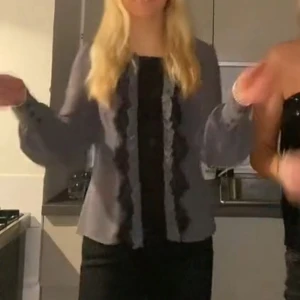 Grå blus med volangdetaljer - Säljer en grå blus med långa ärmar och knappar i ryggen. Blusen har två svarta volangdetaljer framtill som ger en snygg kontrast. Ärmarna är lite puffiga och avslutas med breda muddar. Perfekt för dig som gillar en stilren men ändå unik look.