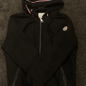 Svart vindjacka från Moncler med huva - Svart vindjacka från Moncler i storlek S. Jackan har huva med snörning och snygga rödvita detaljer, samt dragkedja framtill och på fickorna. Moncler-logga på ärmen. Perfekt för dig som gillar stilrena och sportiga plagg.