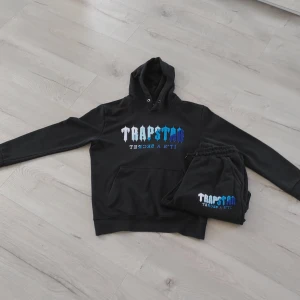Svart Trapstar hoodie med blått tryck - Säljer en svart hoodie från Trapstar med stort tryck i vitt och blått på bröstet där det står 'Trapstar It's a secret'. Hoodien har huva med snörning och en klassisk känguruficka framtill. Perfekt för dig som gillar streetwear och vill sticka ut.