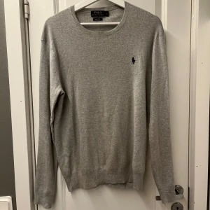 Grå tröja från Polo Ralph Lauren - Klassisk grå tröja från Polo Ralph Lauren med rund halsringning och diskret broderad logga på bröstet. Långärmad och i mjuk bomull, perfekt för lager-på-lager. Enkel och stilren design som funkar till allt. Den ända defekten är att den är lite  lite solblekt runt halsen.