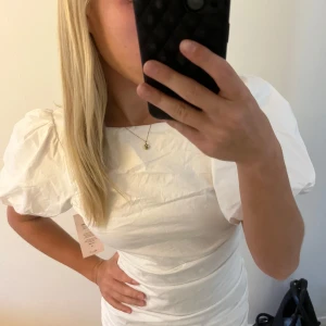 Vit puffärm kortklänning - Superfin vit kort klänning med puffärmar och rund halsringning. Sitter figurnära med snygga rynkade detaljer längs sidorna. Perfekt för dig som vill sticka ut med en stilren och trendig look. Materialet känns lätt och passar bra till varmare dagar. Helt ny med lapp kvar!