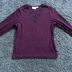 Mörklila långärmad topp med spets - Snygg mörklila långärmad topp med v-ringning och dekorativ spetsdetalj framtill. Toppens material är mjukt och stretchigt och tunn. Storlek 146/152