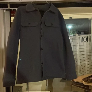 Mörkblå overshirt med fickor - Stilren mörkblå overshirt med klassisk krage och två bröstfickor med knappar. Skjortan har långa ärmar och knäppning framtill. Perfekt att slänga över en t-shirt för en chill look.