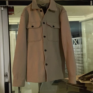 Beige overshirt med fickor och knappar - Säljer en stilren beige overshirt med klassisk krage och svarta knappar framtill. Jackan har två stora bröstfickor med lock och knapp, samt långa ärmar med knapp vid ärmslut. Perfekt för lager-på-lager och enkel att matcha.