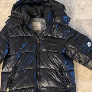 Moncler Maya  - Snygg svart pufferjacka från Moncler med glansig finish och huva. Jackan har dragkedja framtill, två fickor med dragkedja och klassisk Moncler-logga på ärmen. Insidan har ett coolt serietidningsmärke. Perfekt för kalla dagar och riktigt street. Jackan är inprincip ny då jag köpte jackan sent på vintersäsongen. 100% äkta och kvitto finns. Jackan är i storlek 3 vilket motsvarar S-M 