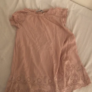 Ljusrosa kortklänning från 157 - Supersöt ljusrosa kortklänning från 157 med broderade detaljer och hålmönster på ärmar och nederkant. Klänningen har korta ärmar och en avslappnad passform, perfekt för varma dagar. Mjuk och skön i bomull.