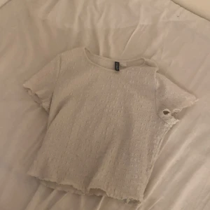 Vit strukturerad topp från H&M Divided - Säljer en vit kortärmad topp från H&M Divided i storlek M. Toppen har en strukturerad, lite ribbad yta och råa kanter vid ärmslut och nederkant. Enkel och stilren modell som passar perfekt till jeans eller kjol.